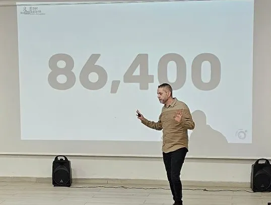 עזר סלם מרצה על במה, מול מסך שעליו המספר 86,400